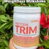 A tub of Vita Optimal Protein Trim in Vanilla Milk Shake flavour, 640g, held in front of a hibiscus plant